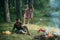 Camping concept. Thoughtful man looking at flames and drinking coffee. Guy sitting on sunny meadow. Two barefoot girls