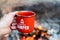 Camping lifestyle concept. Hands holding red enamel mug near a campfire