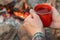 Camping lifestyle concept. Hands holding red enamel mug near a campfire