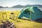 Camping tent with extinguished bonfire in the green field meadow, Lake and mountain background. Picnic and travel concept. Nature
