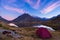 Camping with tent near high altitude lake on the Alps. Reflection of snowcapped mountain range and scenic colorful sky at sunset.