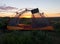 Camping tent with solar charger in quiet grassy meadow while the sunset peers through mesh screen. A peaceful campsite for the eve