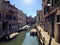 A canal in Venice, Italy on a quiet summer morning.  Boats line the side of the canal with a few people walking beside the canal