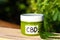 Cannabis medical products in jar with the inscription CBD with green leaves, CBD lotion on blurred background of hemp bush