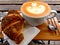 Cappuccino with froth and fresh croissant on the side on bamboo tray