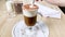 Cappuccino, latte with froth milk in cup. Hand stirs spoon of coffee in tall glass, scoops up whipped cream, sprinkled with ground