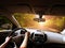 Car dashboard with driver`s hands on the steering wheel and rear view mirrors on a road in motion with trees against sky with