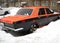 A car parked in the courtyard of apartment buildings is littered with snow. The machine is painted in orange and black. A sickle