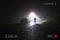 A car parked on the side of the road, underneath a street light, with a hooded figure, on a rural, country road. On a foggy winter