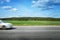 The car speeds along a country road, surrounded by forest and blue sky