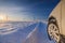 Car tires on winter road covered with snow. Vehicle on snowy country road with wind turbine in the background. Snow covered field