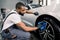 Car washing and detailing photo. African man worker in protective overalls and rubber gloves, washing car alloy wheel on