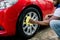 Car washing and detailing photo, car wash outdoors. Cropped image of young African man, washing red car alloy wheel on a