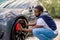 Car washing on open air. Young hipster African bearded man cleaning a wheel, car rims of modern luxury gray electric car