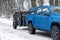 Caravan or mini camper towed by blue muscle car in a white snowed forest. Family journey or away time in a wild. Caravan