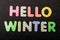 Card with Hello Winter words made from mixed vivid colored wooden letters on a textured dark black textile material that can be