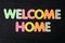 Card with Welcome Home words made from mixed vivid colored wooden letters on a textured dark black textile material that can be