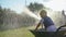 Carefree childhood, happy wet girl in wheelbarrow with garden hose watering green grass in park at during summer holiday