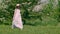 Carefree girl in hat and long white dress walking on flowering meadow in orchard. Wind blowing hat from walking teen