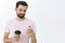 Carefree happy and calm attractive european man with beard and cool hairstyle holding smartphone and paper cup of coffee