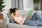 Carefree korean lady lying on couch in living room, reading book while resting at home at weekend, copy space