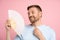 Carefree man using paper fan at summer hot weather enjoying fresh air isolated on studio pink