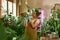 Careful female gardener wearing gloves smiling and looking at potted plant while posing indoors