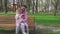 A caregiver and a girl are sitting on a park bench and are busy on a smartphone.