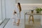 Caring mother combs hair of her small daughter wear white bathrobes pose against kitchen interior stand bare feet at floor.