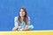 Casual adult woman lean on yellow wall holding a mobile phone while looking away outdoors against a blue background