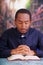 Catholic priest wearing traditional clerical collar shirt sitting with folded hands holding rosary while praying and