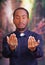 Catholic priest wearing traditional clerical collar shirt standing facing camera, holding hands out with rosary cross