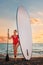 Caucasian adult plump woman in a red swimsuit holding a sup board and paddle. Sunset sky on the background. Vertical