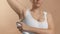 Caucasian anonymous girl in white top with a hand spray deodorant on her underarm. Studio photo image on beige