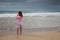 Caucasian baby girl in pink stands on the sea coast