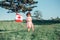 Caucasian baby toddler girl standing on green grass in park outside and holding waving large Canadian flag.