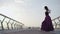 Caucasian ballerina jumping up at sunrise and standing in ballet position on bridge. Side view wide shot of brunette