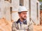 Caucasian bearded civil engineer or construction worker in white hardhat stands cross handed opposite construction site