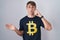 Caucasian blond man wearing bitcoin t shirt confused and annoyed with open palm showing copy space and pointing finger to forehead