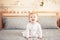 Caucasian blonde baby girl in white onesie sitting on bed in bedroom