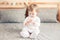 Caucasian blonde baby girl in white onesie sitting on bed in bedroom