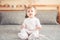 Caucasian blonde baby girl in white onesie sitting on bed in bedroom