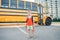 Caucasian boy student with backpack near yellow bus on first September day. Child kid eating apple fruit at school yard outdoor.