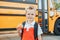 Caucasian boy student with backpack near yellow bus on first September day. Child kid eating apple fruit at school yard outdoors.