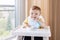 Caucasian child boy with dirty messy face sitting in high chair eating apple puree with spoon