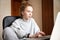 Caucasian cute red-haired teenage girl in a grey hoodie and pants, sitting at a table in front of a laptop indoors. Home