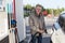 Caucasian elderly man is on gas station with fuelling nozzle inserted in car filler well