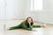 Caucasian female model demonstrates side split, practices yoga, dressed in green sweatsuit, white sneakers, poses in empty hall on