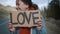 Caucasian girl hipster in the daytime stand on side of two lane highway hitchhiking while holding cardboard sign with