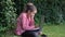 Caucasian girl schoolgirl sits in a park on a green lawn with a laptop on her feet watching a video and makes notes with